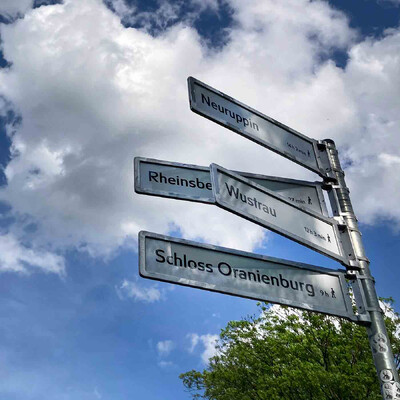 Aluminiummast mit vier Schildern vor einem blauen Himmel mit weißen Wolken: Neuruppin 14 Stunden 2 Minuten, verdeckt vom nächsten Schild: Rheinsbe… , Wustrau 12 Stunden 3 Minuten, Schloss Oranienburg 9 Stunden